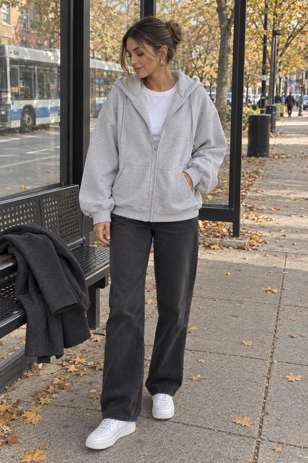 A woman without sunglasses wearing an oversized grey zip hoodie, dark trousers and white sneakers, with her charcoal coat on the bench beside her