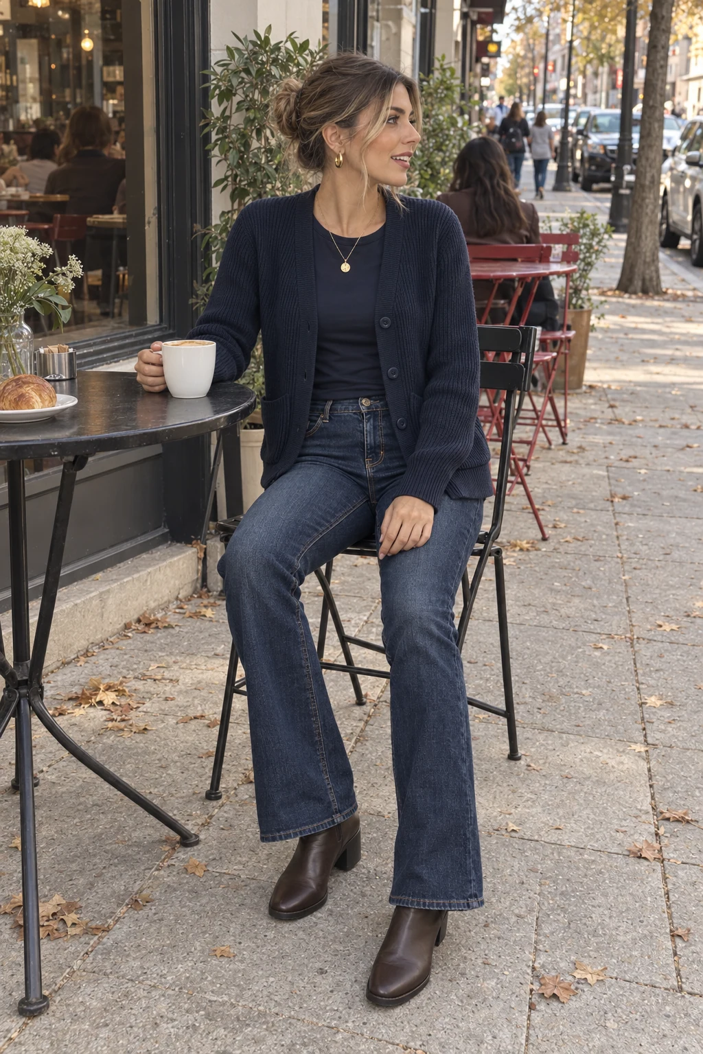 A woman seated at an outdoor cafe without sunglasses in a navy pocket cardigan, dark indigo bootcut jeans and dark ankle boots