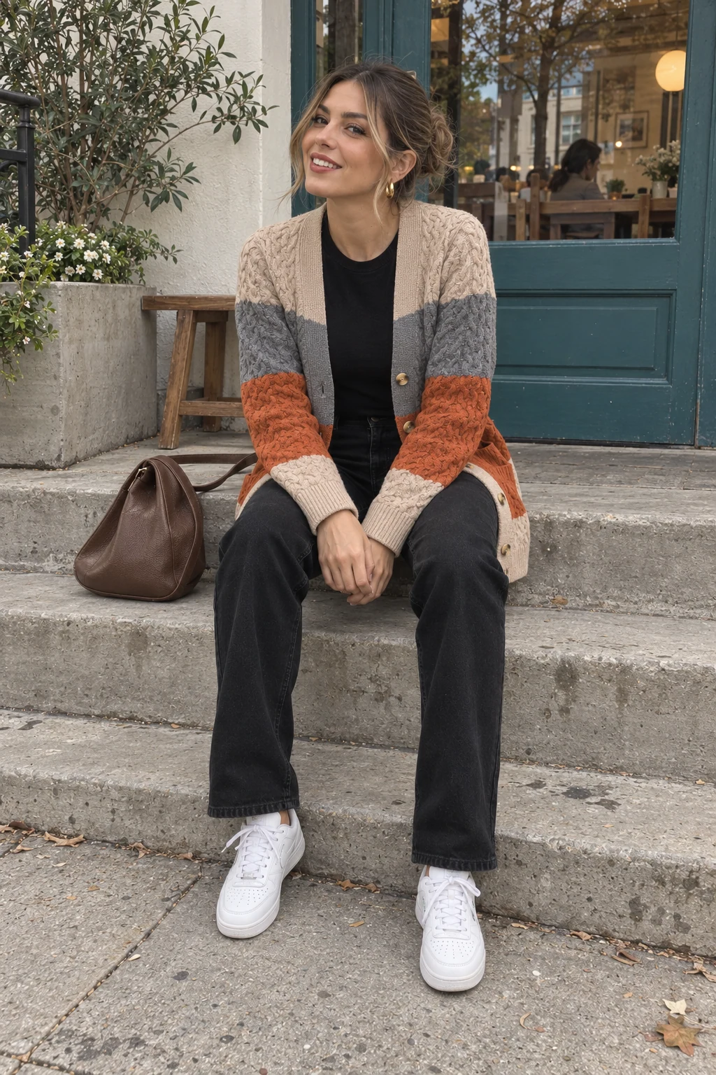 A woman sitting on neighborhood cafe steps without sunglasses in an autumn colorblock cardigan, black straight-leg jeans and white sneakers